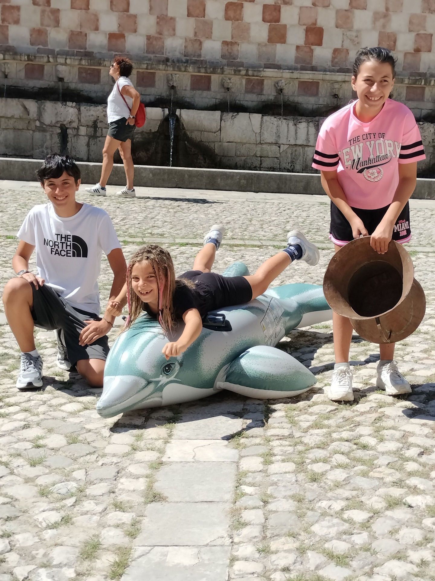 Tre bambini sorridenti posano all’aperto su una scultura a forma di delfino azzurro, con sfondo in mattoni.