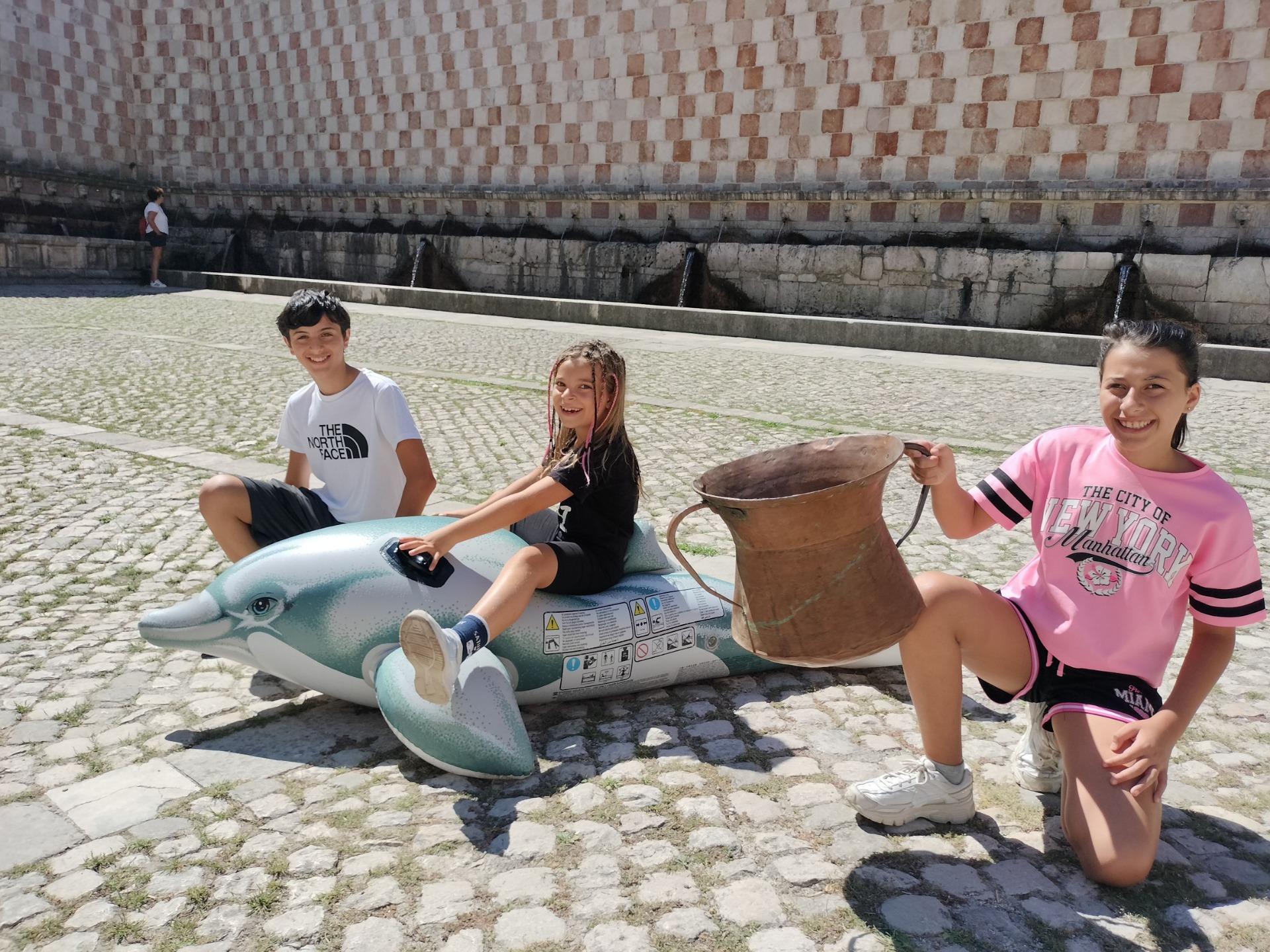 Tre bambini alle 99 cannelle di L'Aquila, con un delfino gonfiabile e un'antica conca.