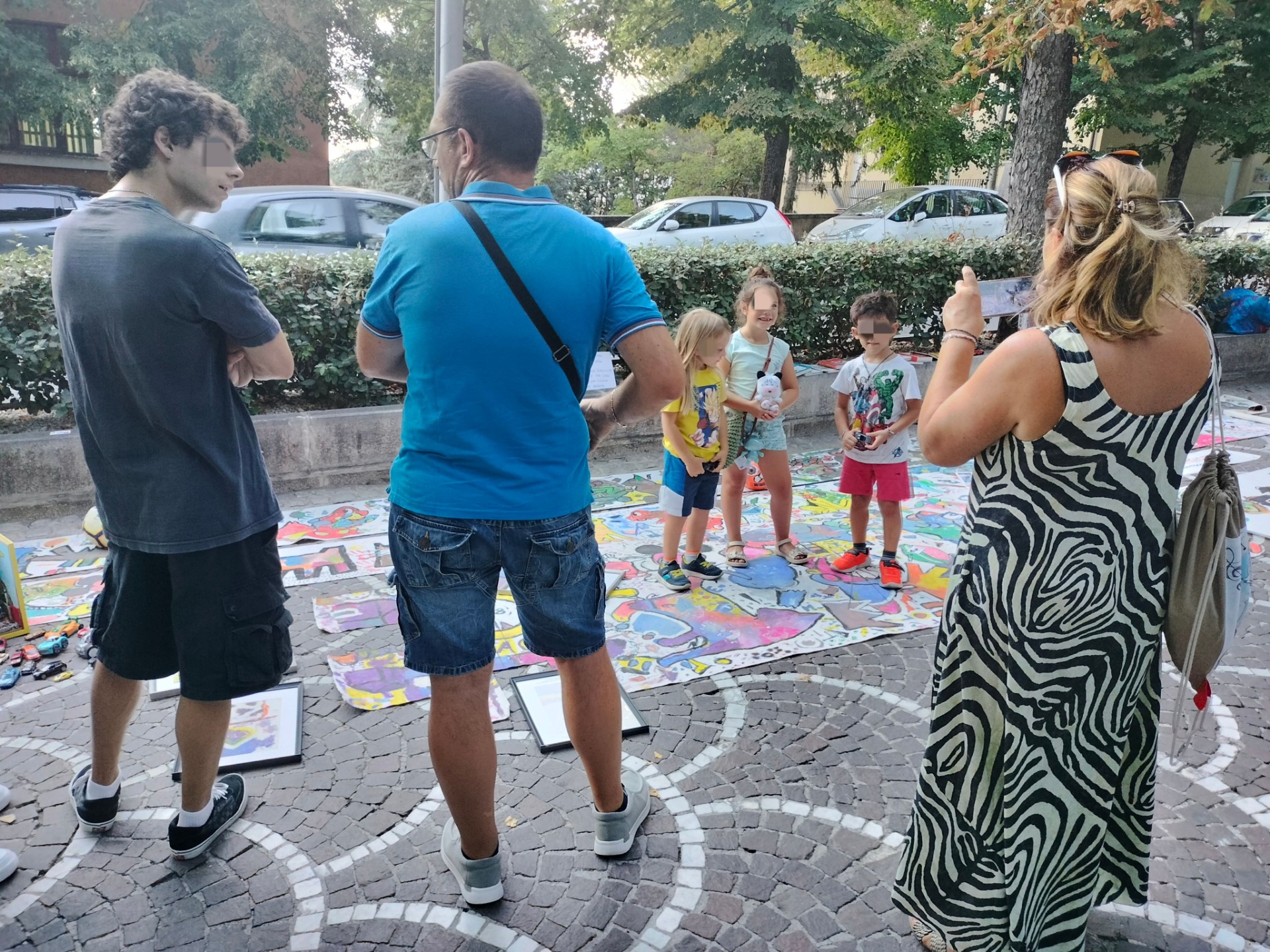 Adulti e bambini osservano disegni colorati su pavimento, in un parco