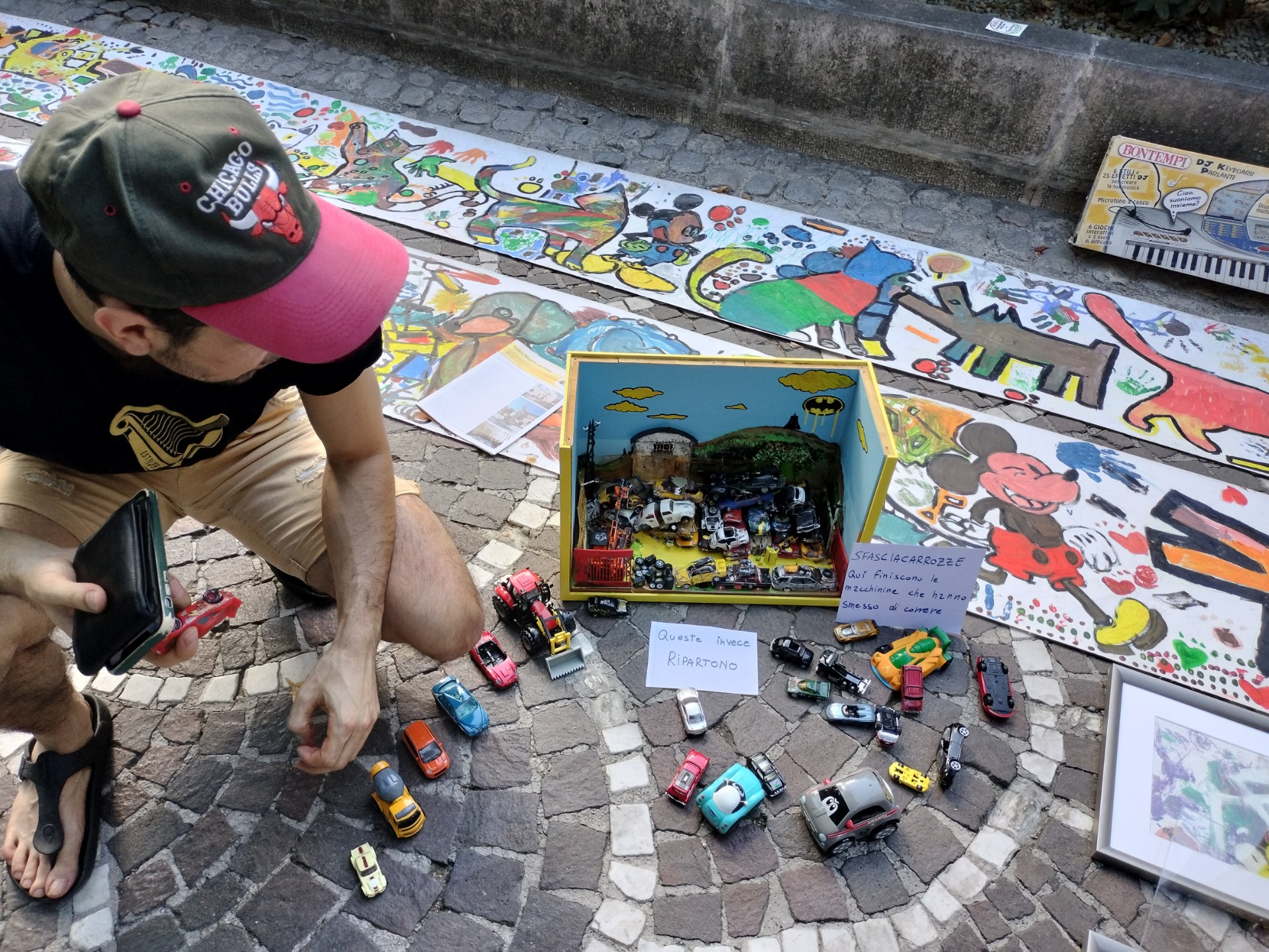 Uomo con cappellino rosa e verde si china su modellini di auto esposti su strada con murales.