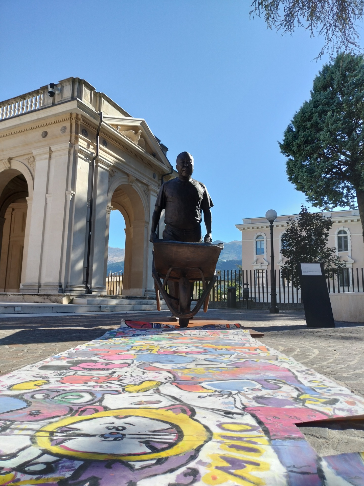 Statua in bronzo su basamento, davanti a edificio neoclassico, con murale colorato a terra.