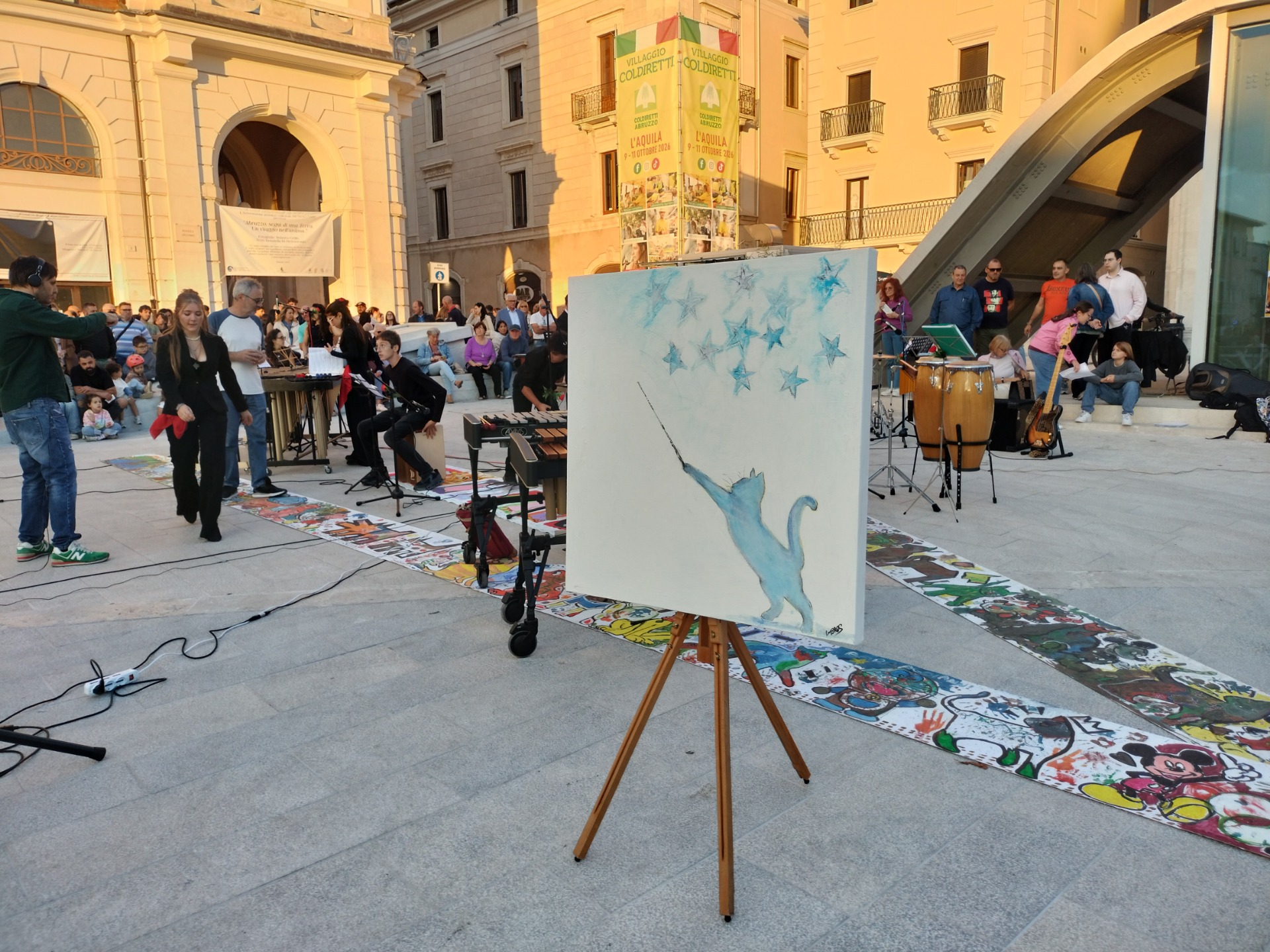 Piazza con musicisti e pubblico; cavalletto con grande dipinto blu di stelle e gatto con bacchetta da direttore d'orchestra
.