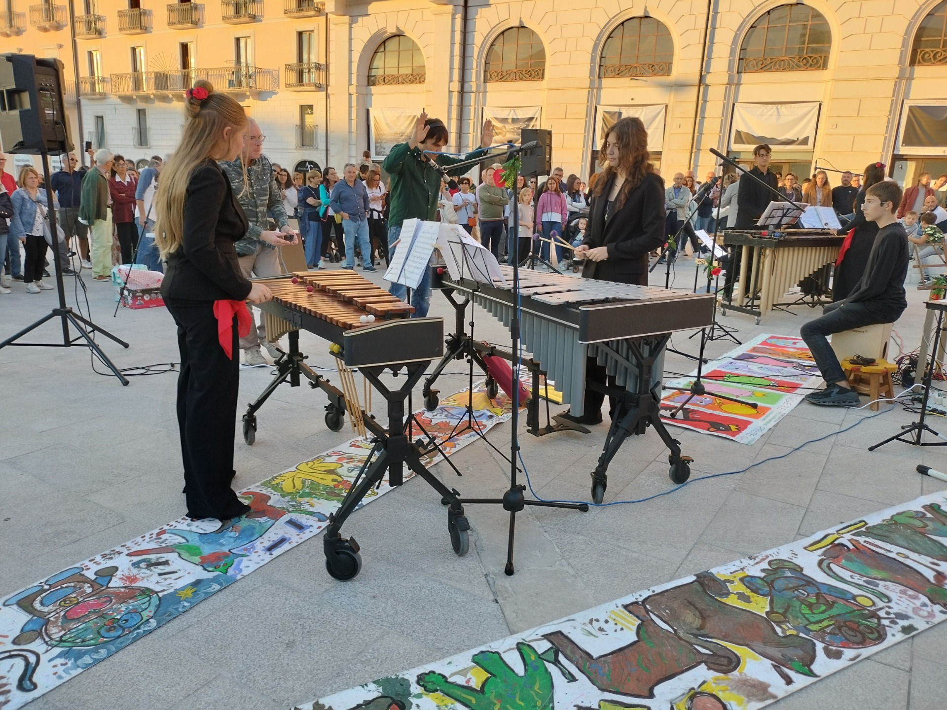 Gruppo musicale con due marimbisti e pubblico in piazza, strumenti a percussione su tappeti colorati dai bambini