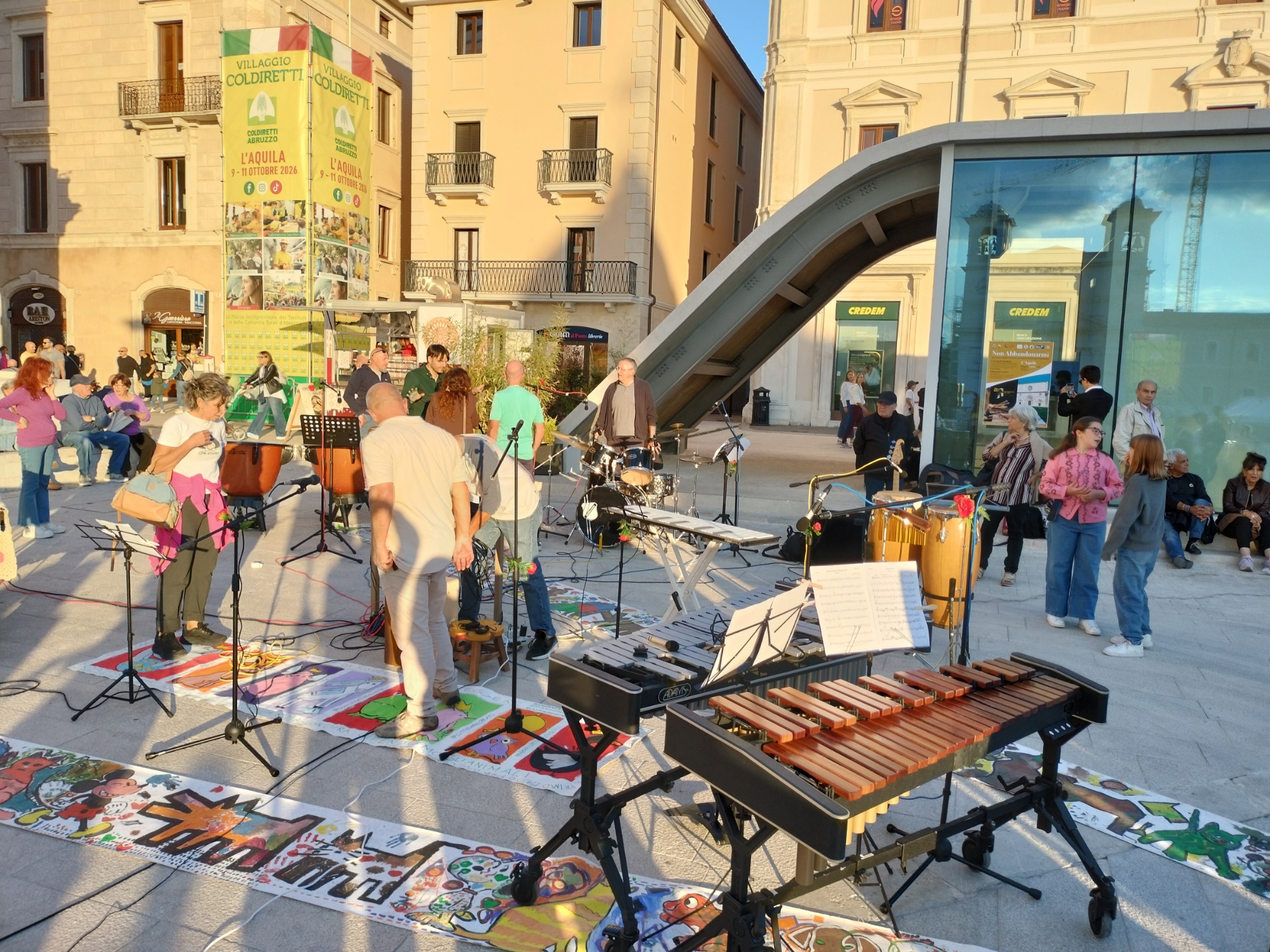 Gruppo musicale su palco con strumenti a percussione e xilofono, pubblico in piazza davanti a edifici storici.