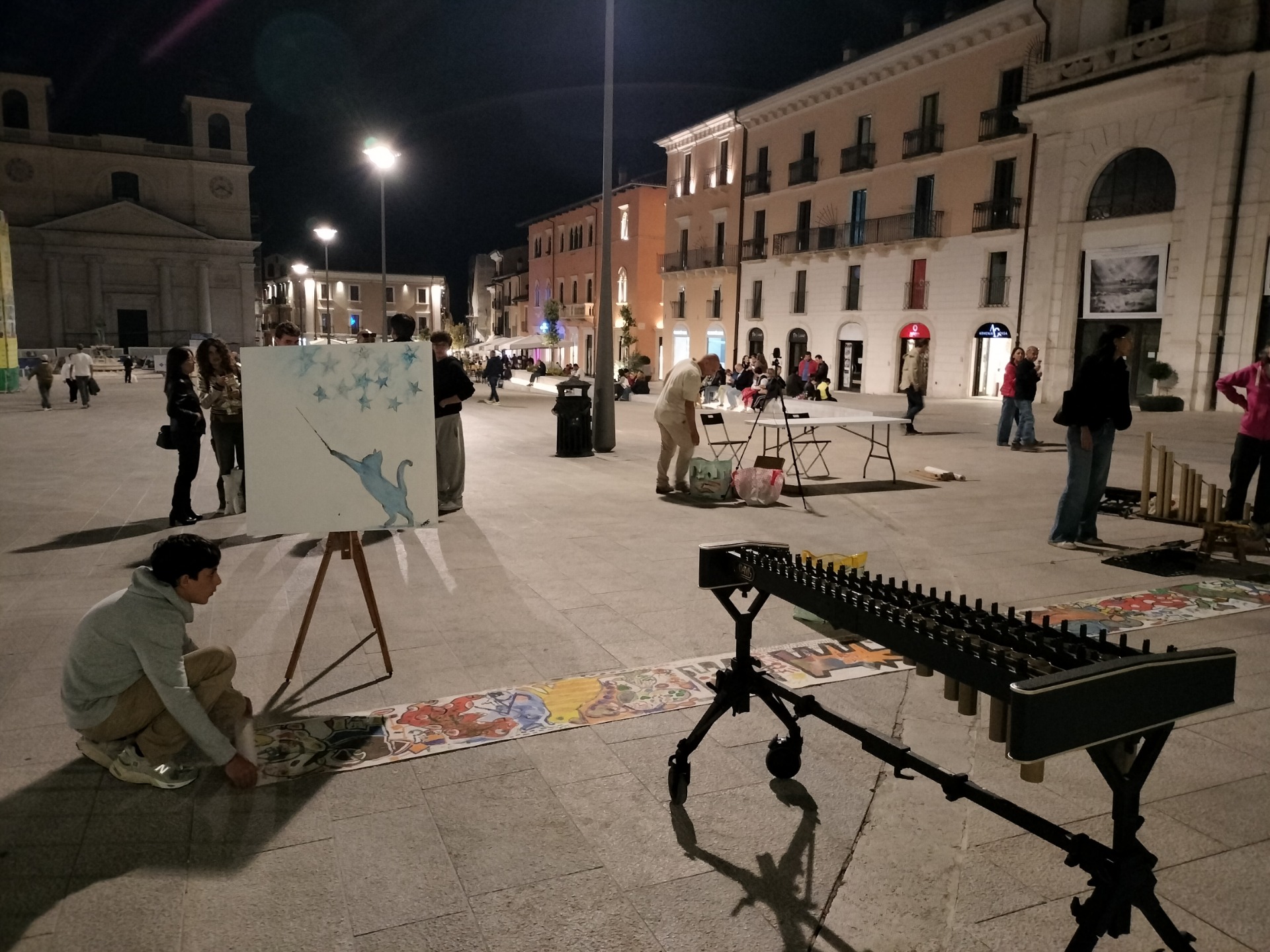 Piazza notturna con artisti, cavalletto e dipinto. Strumenti e persone davanti a edifici illuminati.
