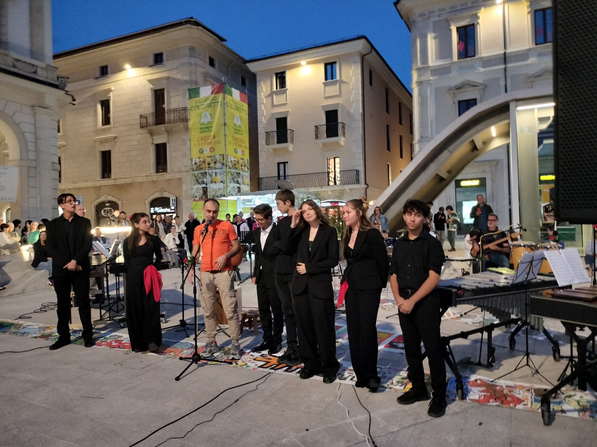 Gruppo di giovani in abiti scuri su strada coloroata  con strumenti, durante concerto serale in piazza illuminata.