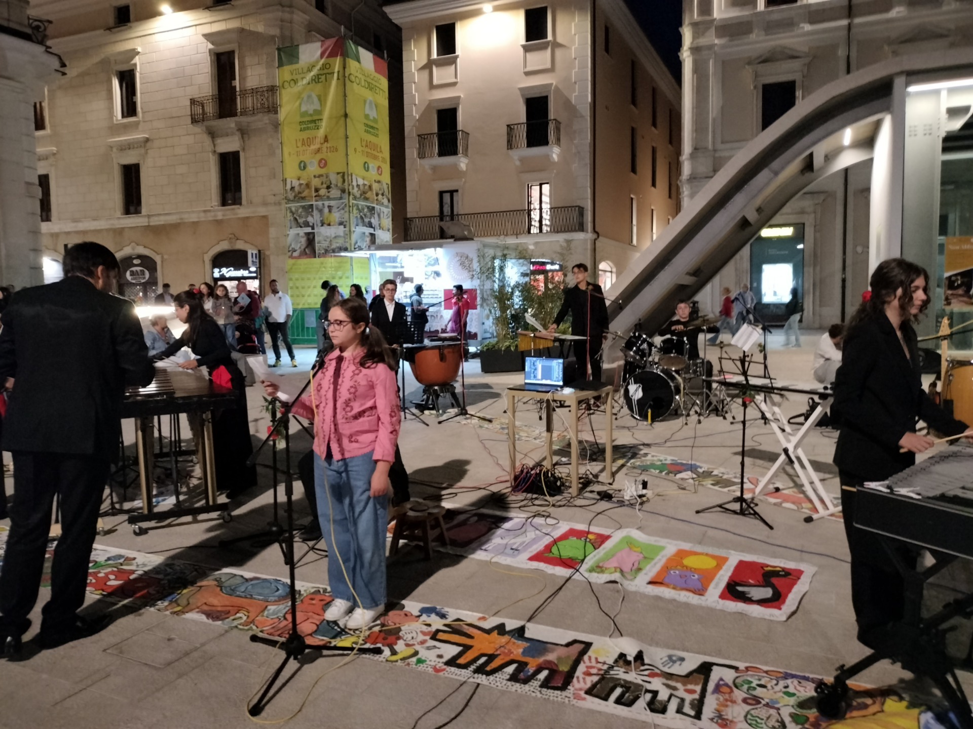 Concerto in piazza con musicisti su palco, strumenti e cartelloni colorati tra edifici illuminati serali.