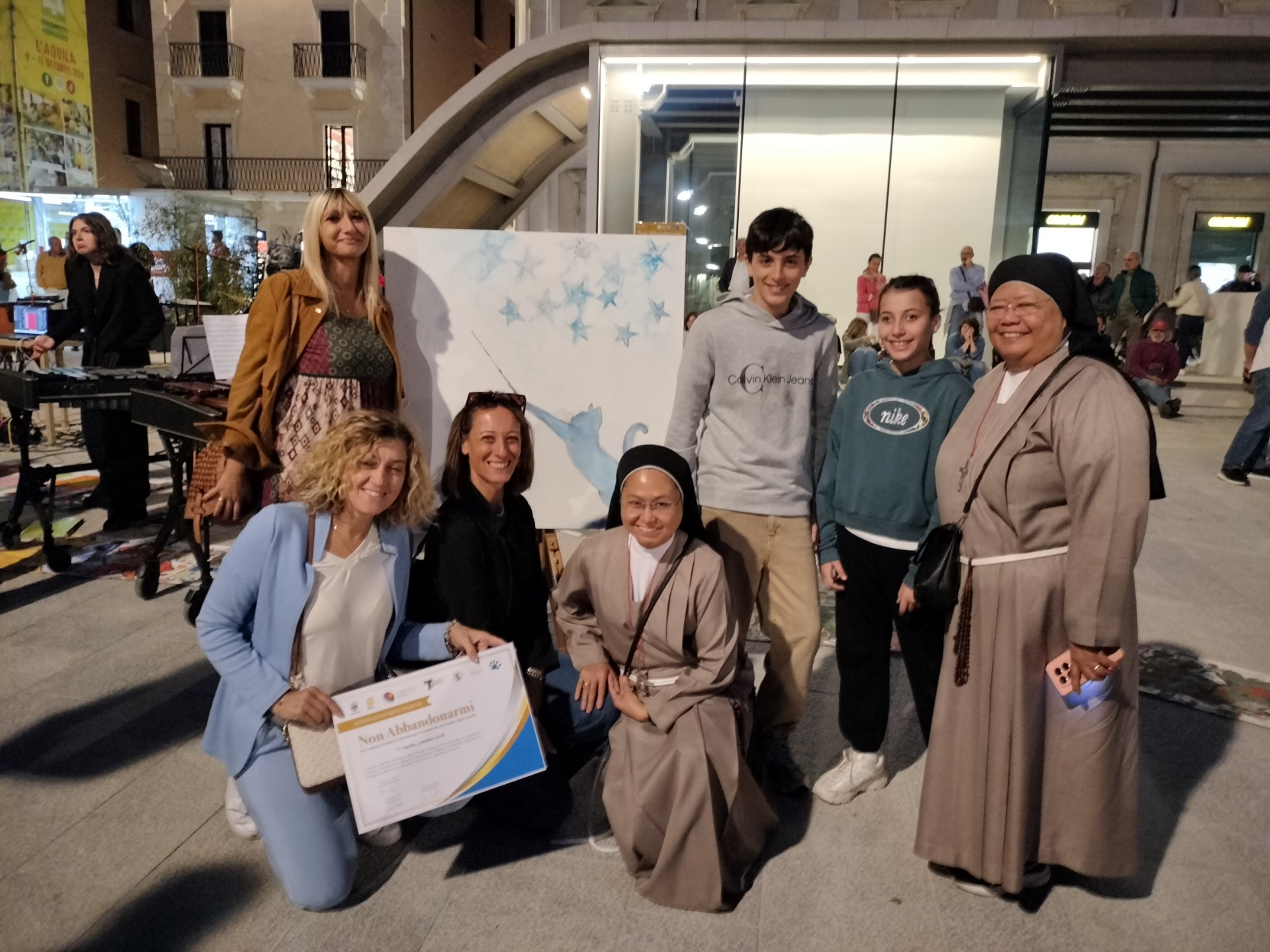 Gruppo di persone sorridenti in atrio, alcune sedute, una tiene un attestato, sullo sfondo pannello con stelle.