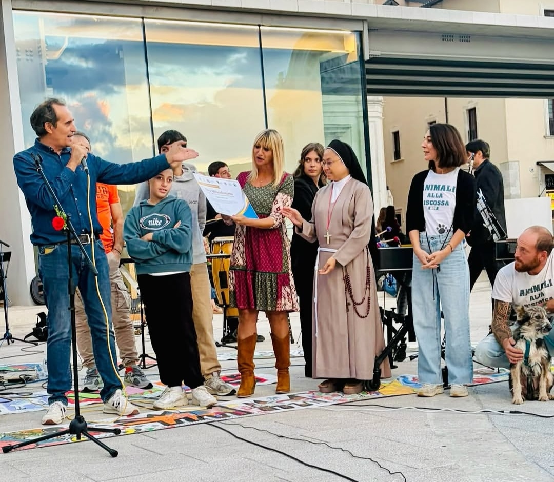 Gruppo in piedi su palco all’aperto, una donna riceve un attestato mentre altri ascoltano e applaudono.